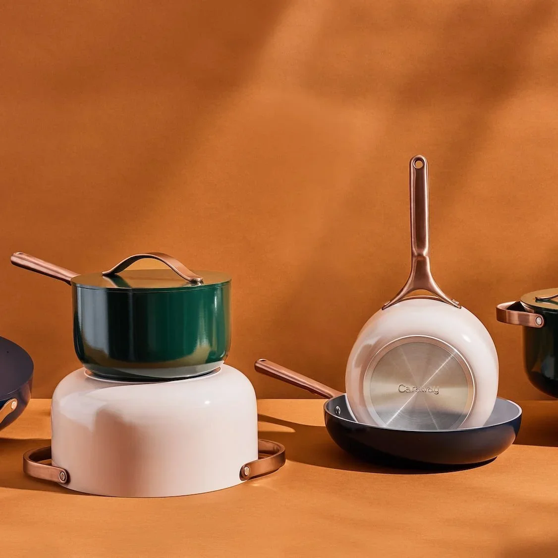 Set of colorful pots and pans displayed on a wooden surface against a warm brown background, including a white pot, a green pot, and a white frying pan with a copper handle.