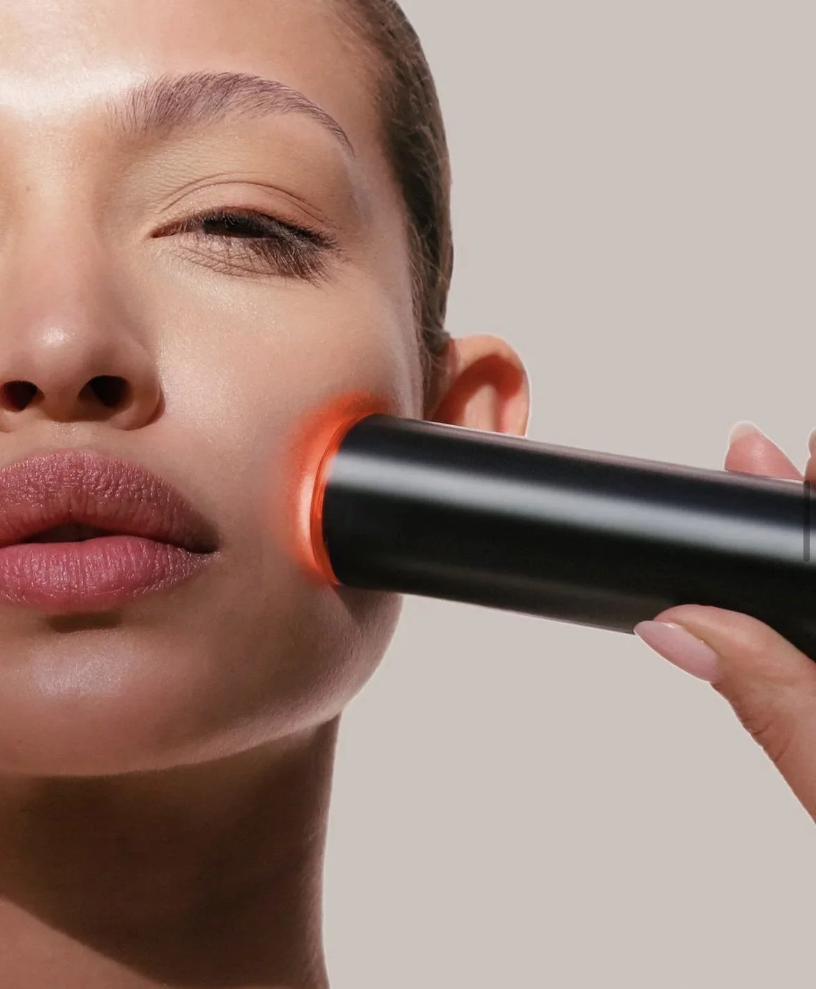 Close-up of a woman with her eye closed, receiving a facial treatment from a black handheld device emitting red light on her cheek.