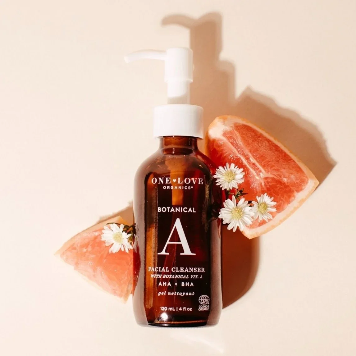 A brown bottle of One Love Organics Botanical A Facial Cleanser with a white pump, surrounded by grapefruit slices and small white flowers, placed on a light surface.