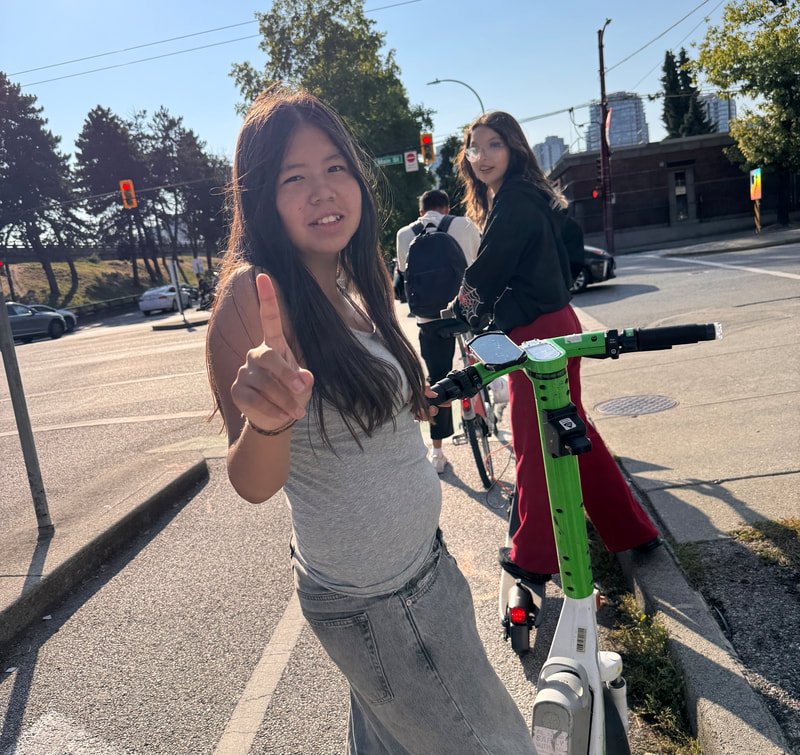 Young woman with long dark hair wearing a gray t-shirt and gray jeans gesturing with her index finger, standing next to a green and white electric scooter on a city street on a sunny day.