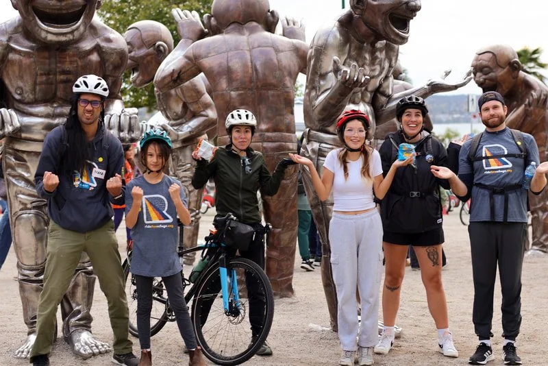 A group of six diverse cyclists, including children and adults, posing in front of large bronze statues of laughing and joyful children during the daytime.
