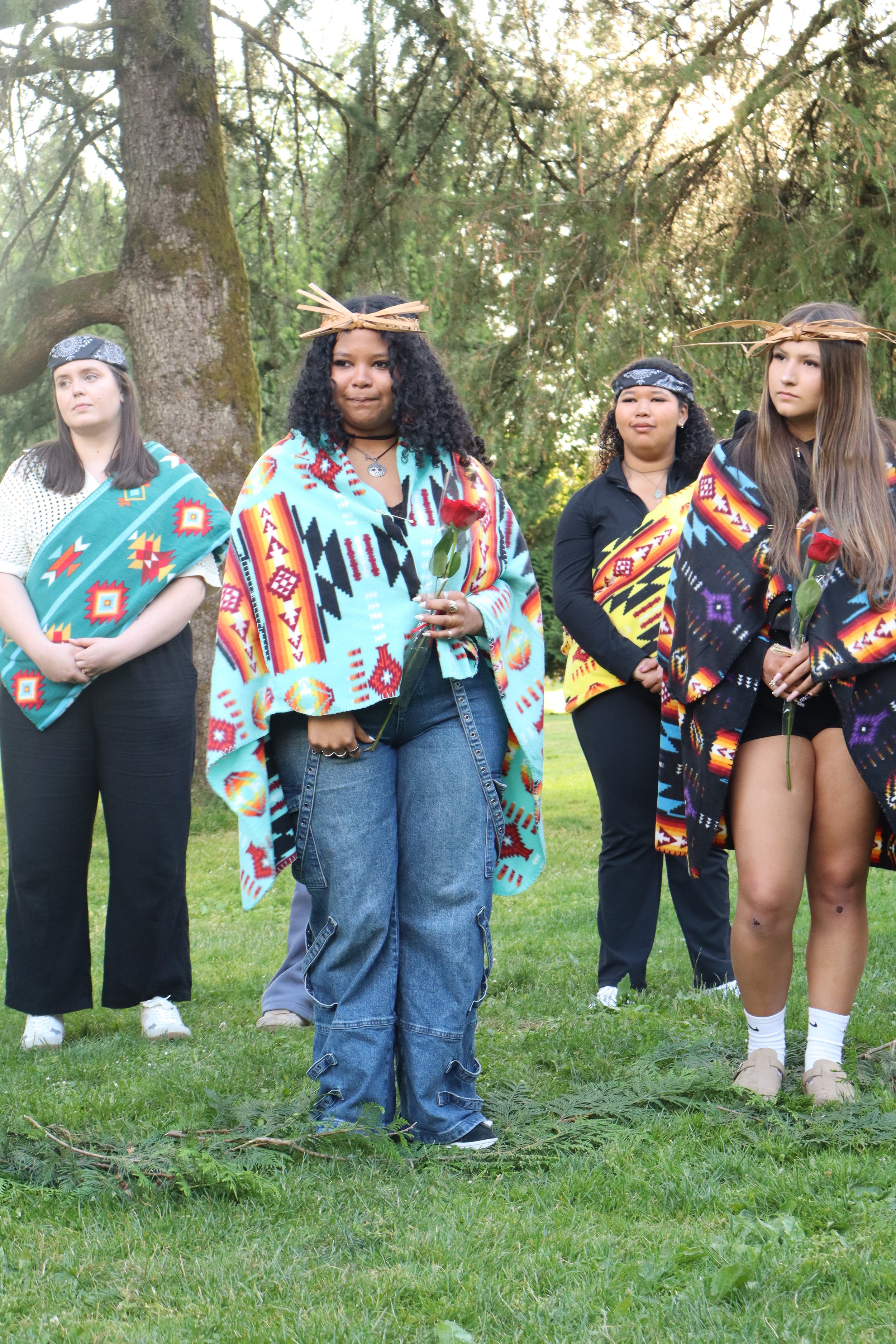 Group of women standing outdoors among trees, dressed in indigenous or traditional clothing, some holding single red roses, some with head wreaths or headbands, on a grassy area in a park or forest.