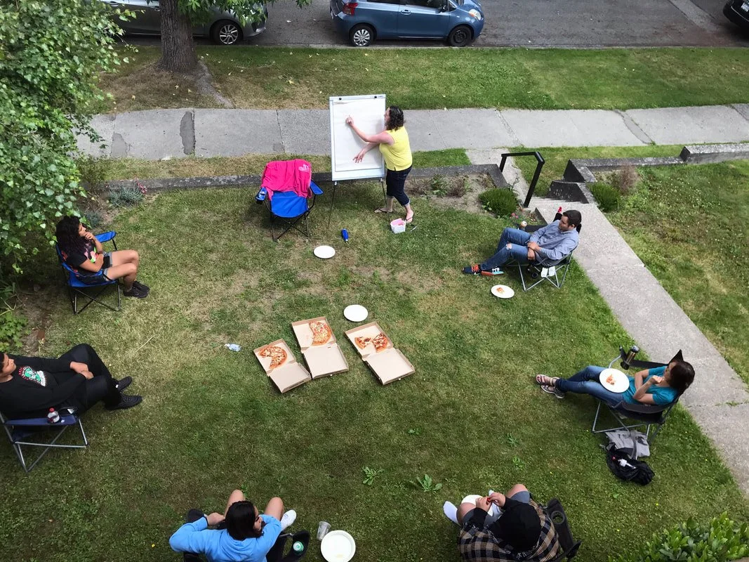 People sitting on lawn chairs around a small gathering, eating pizza, with a person standing at a whiteboard, possibly giving a presentation or teaching, in a backyard with a sidewalk and parked cars visible in the background.