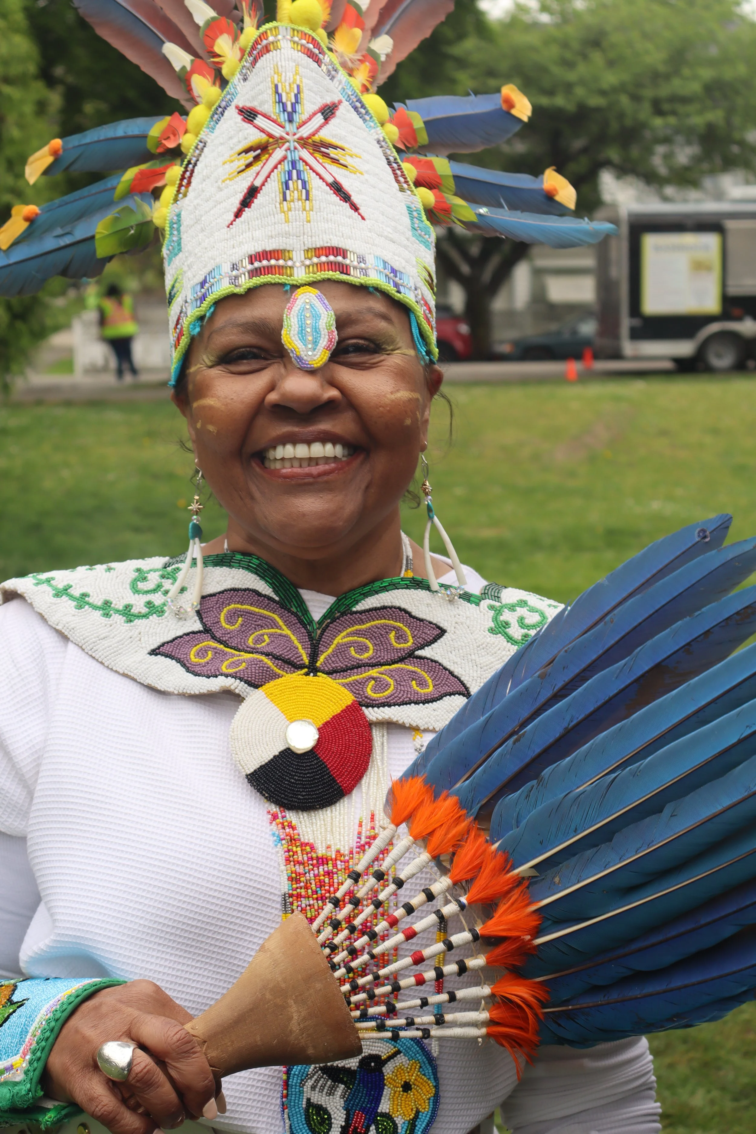 A person dressed in traditional Native American attire, wearing a beaded headdress with colorful feathers, and holding a large feather fan.