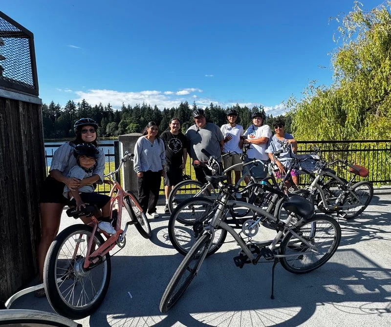 Group of seven people standing outdoors with bicycles on a sunny day, trees in the background.