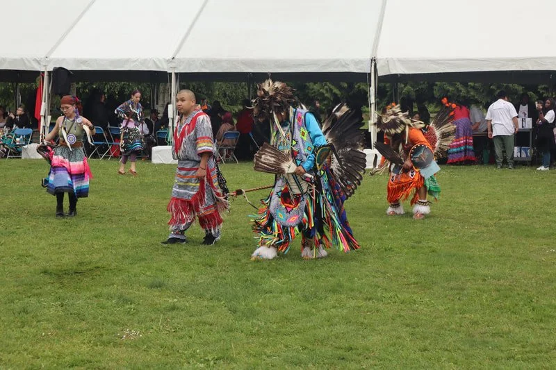 Indigenous people performing traditional dance at an outdoor event under a large white tent, wearing colorful regalia and headdresses.