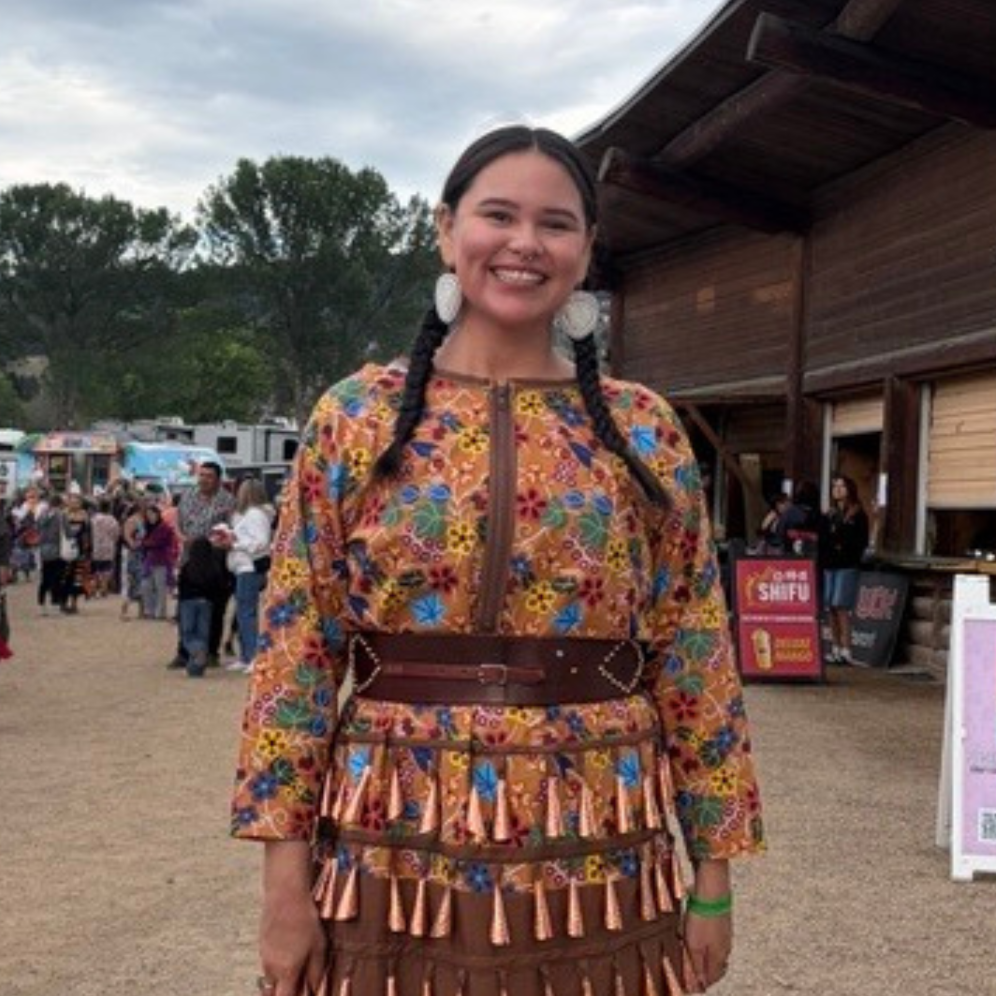A woman with dark hair and large earrings wearing a colorful, floral-patterned dress. She is smiling and standing outdoors at a crowded event, with trees, buildings, and tents visible in the background.
