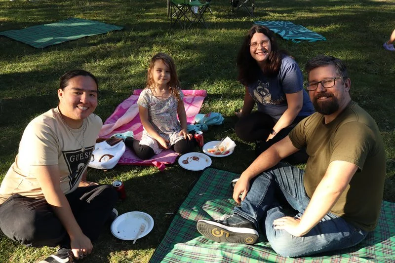 Four people, including a young girl, enjoying a picnic outdoors on a grassy area with blankets and chairs, smiling at the camera.