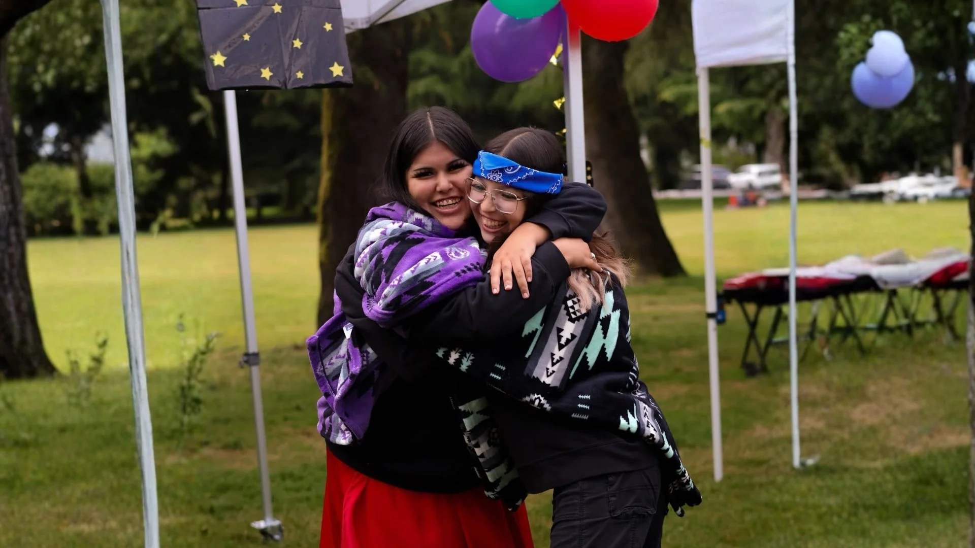 Two women are hugging each other at an outdoor event, smiling. They are surrounded by colorful balloons and a decorated tent in a park with trees and a grassy area.
