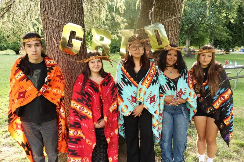 Five teenagers standing outdoors in front of trees, dressed in colorful blankets and wearing woven hats, celebrating a graduation with gold foil balloons spelling 'GRAD' hanging behind them.