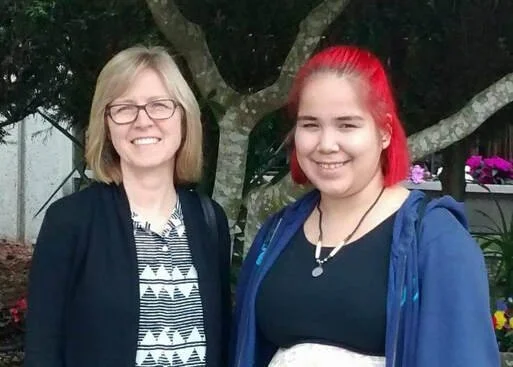 Two women standing outdoors in front of a tree, smiling at the camera, one with blonde hair and glasses, the other with bright red hair.