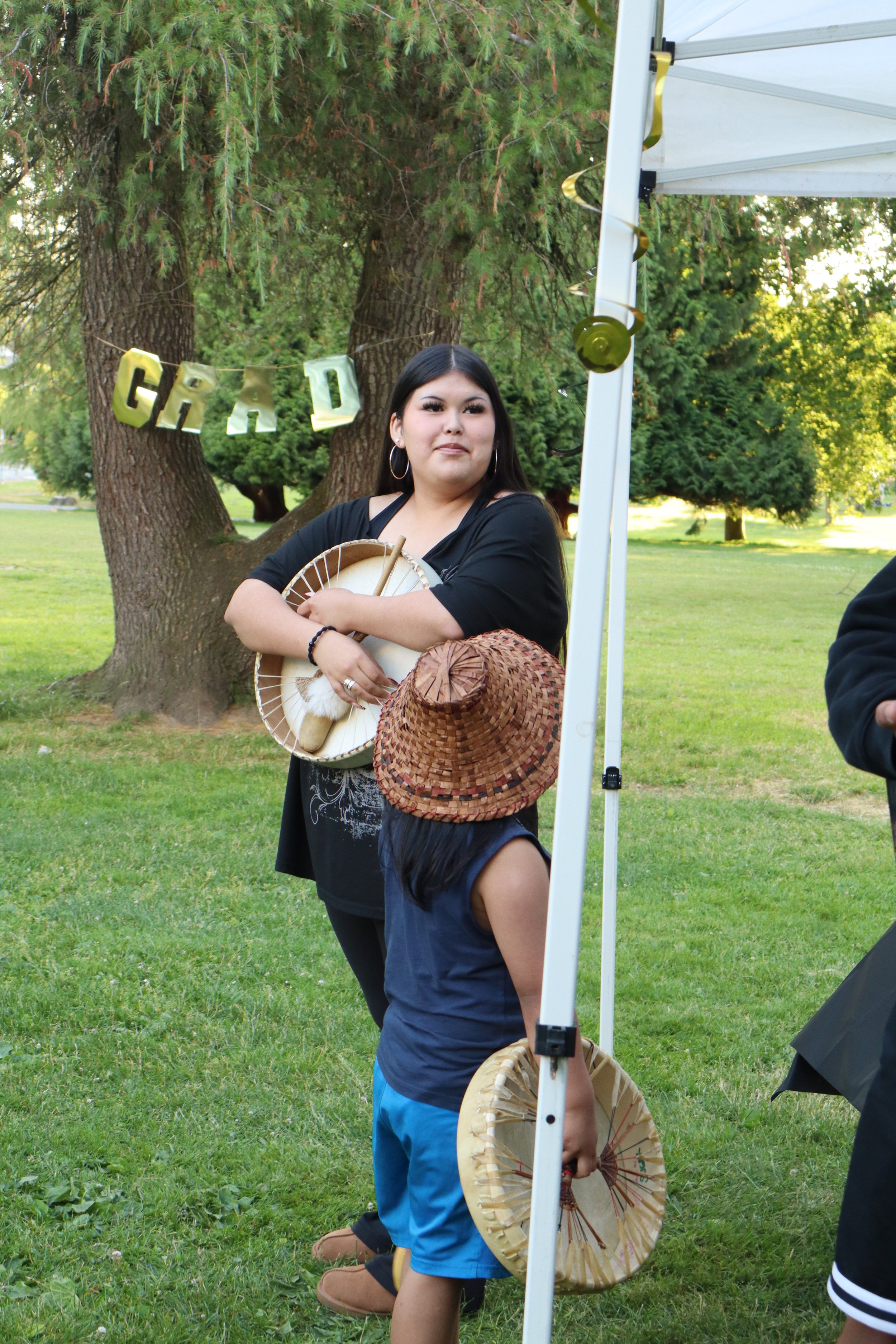 A young woman holding an umbrella in one hand and standing outdoors in a grassy park with large trees behind her. She is wearing a black shirt, earrings, and has dark hair. There is a white canopy tent with gold ribbon decoration nearby and a banner that reads "GRAD" hanging in the background.
