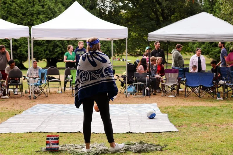 Woman standing on a mat in front of seated crowd under white tents at an outdoor event.