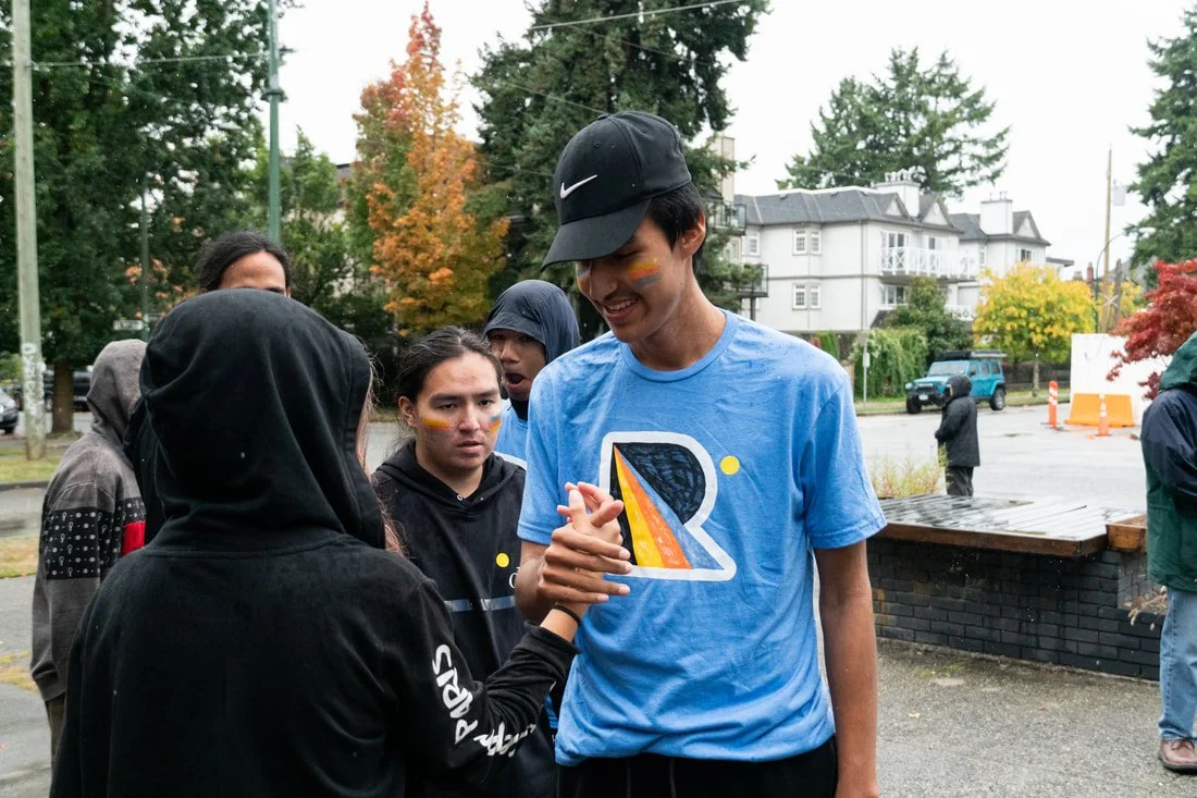 Group of young people outdoors on a rainy day, with some wearing hoodies and jackets, celebrating. A young man in a blue shirt and black cap is smiling and shaking hands with a woman in a black hoodie. The scene is set in a residential neighborhood with trees, houses, and cars in the background.