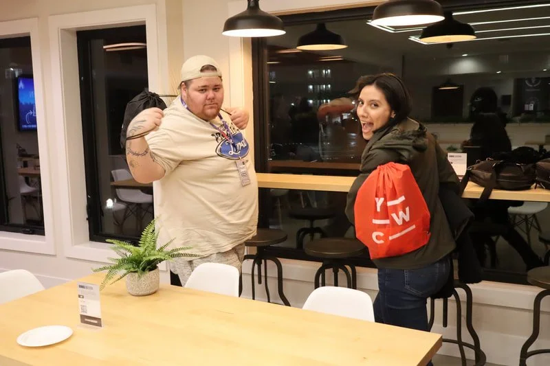 A man and woman standing inside a restaurant or cafe, making playful poses with the man flexing his arm and the woman smiling and turning towards the camera. The woman is wearing a black jacket with a red shopping bag over her shoulder. The man is we