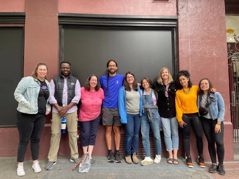 Group of nine diverse people standing outside in front of a dark gray garage door, smiling.