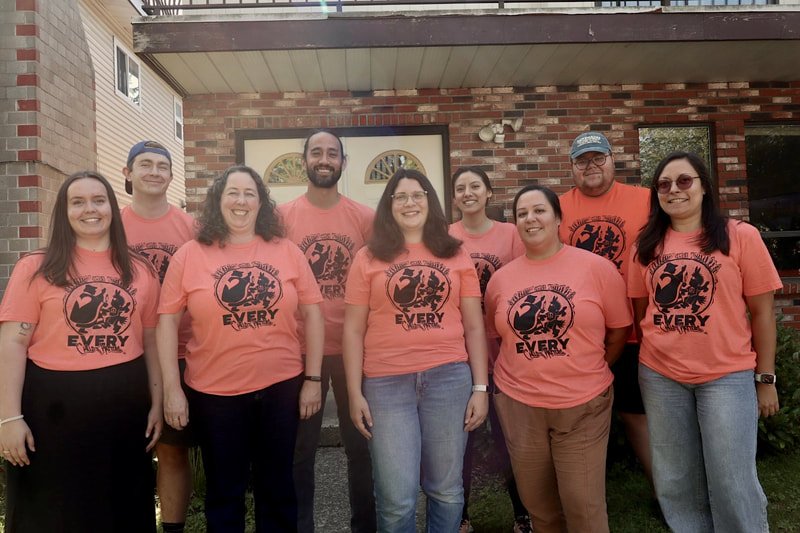 Group of eight people standing outside in front of a brick building, wearing matching pink T-shirts with a black logo and text.