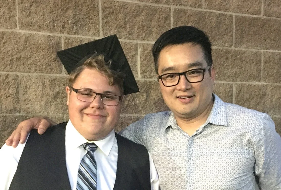 Two men posing for a photo, one wearing a graduation cap and gown, the other in a patterned shirt, both smiling, standing against a brick wall.