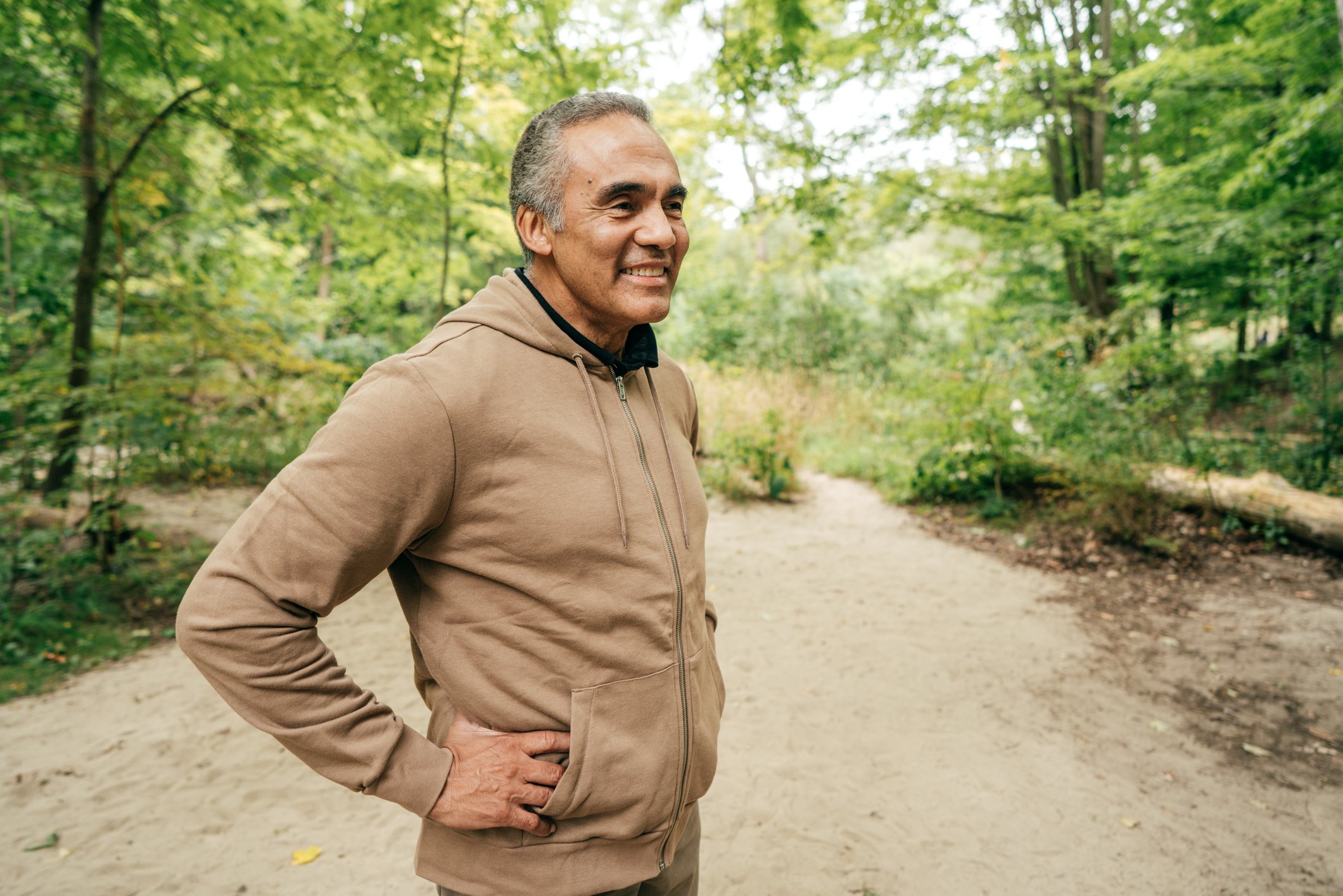 A middle-aged man stands on a forest trail with one hand on his hip and the other resting on his waist, wearing a tan jacket and smiling as he looks to the right, surrounded by green trees.