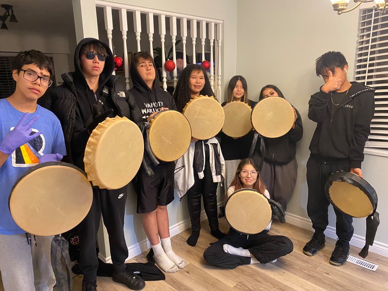 Group of young people holding drums indoors, some wearing gloves, in a room with white walls and windows.