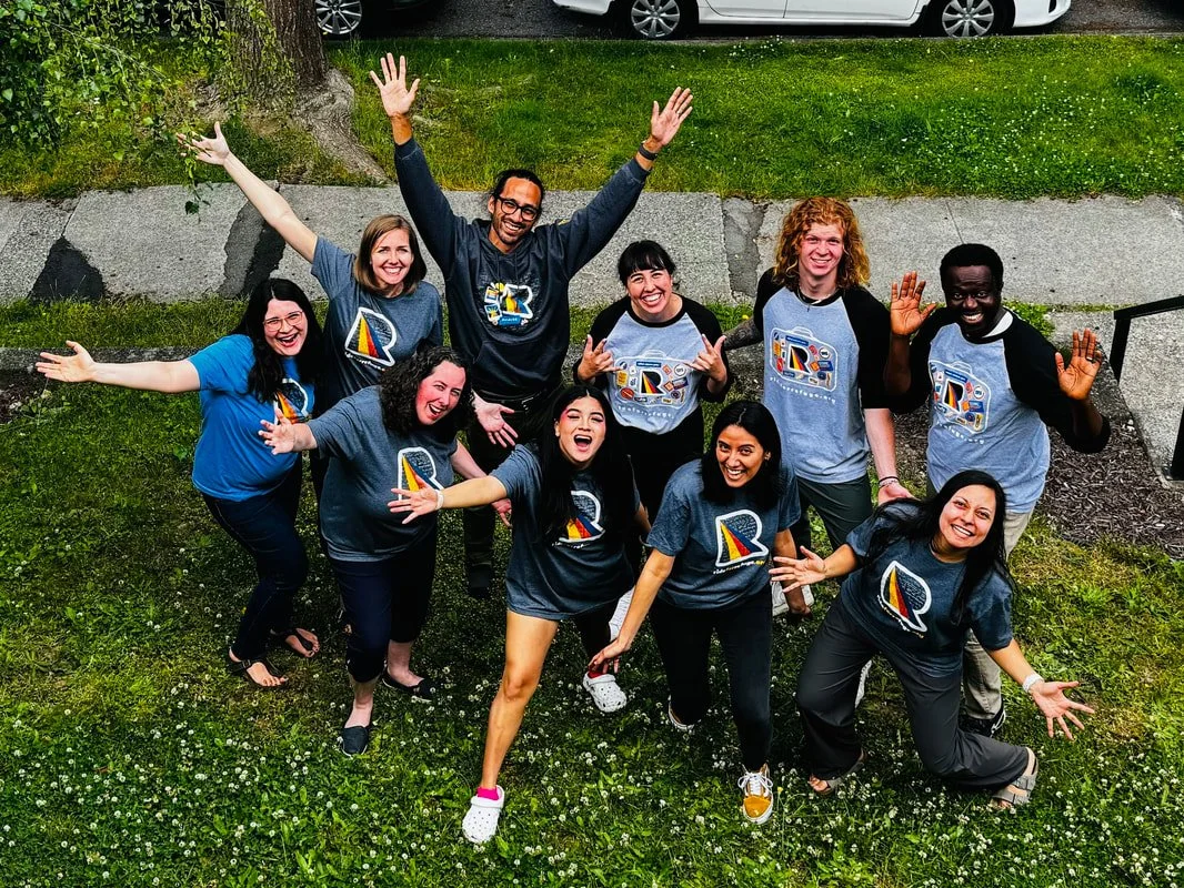 Group of nine people outside on grass, smiling, with some waving, wearing matching shirts with a colorful logo, celebrating or posing cheerfully.