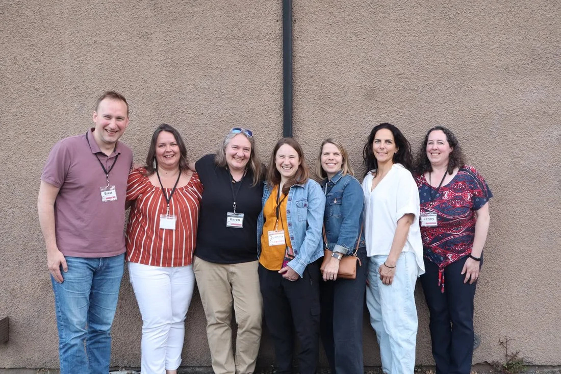 Group of seven smiling adults standing against a tan wall, wearing casual clothing and name tags.