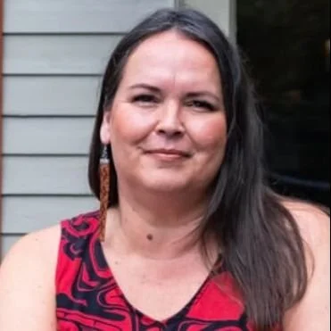 Portrait of a woman with dark hair, wearing a pink and black patterned sleeveless top, standing outside near a house with gray siding.