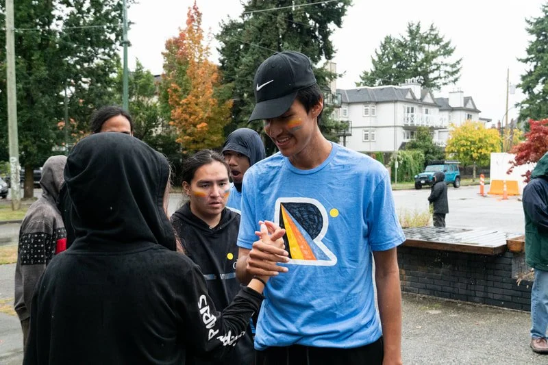Young man in a blue T-shirt, black cap, and with rainbow-colored face paint, smiling as he shakes hands with a young woman in a black hoodie in an outdoor setting with other people around, trees, and houses in the background.