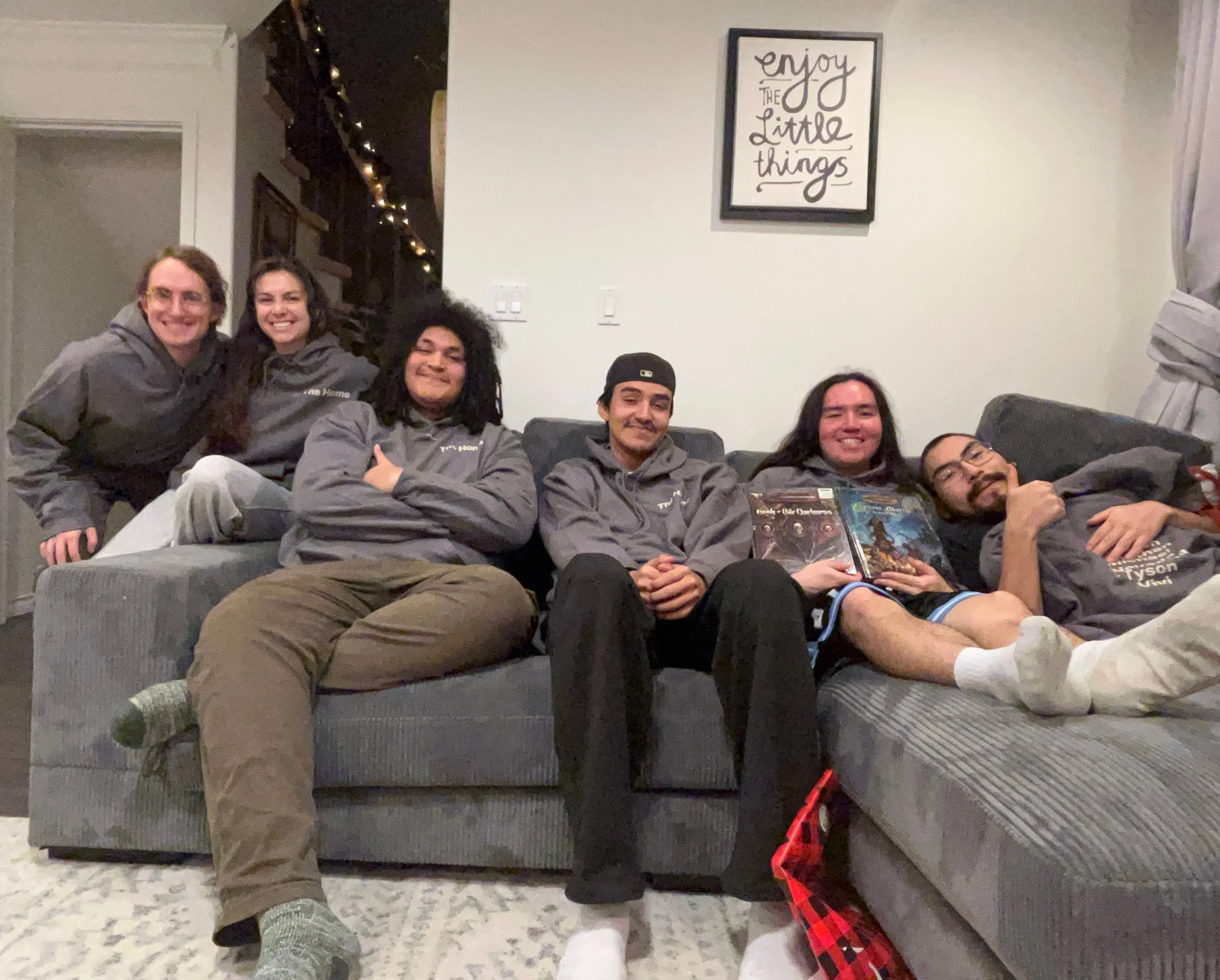 Group of five friends sitting and leaning on a grey couch in a living room, smiling at the camera. They are wearing grey hoodies and casual clothing. Behind them, there is a framed quote on the wall that says 'enjoy the little things.' A person lying on the floor in the foreground is holding a book and has a plaid blanket nearby. The room has a doorway with string lights and a curtain partially visible.
