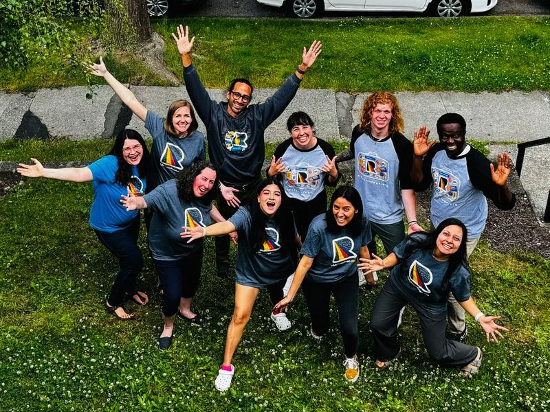 Group of nine smiling people outdoors, raising their hands, wearing matching event T-shirts with a sailboat logo, standing on grass near a sidewalk and parked cars.