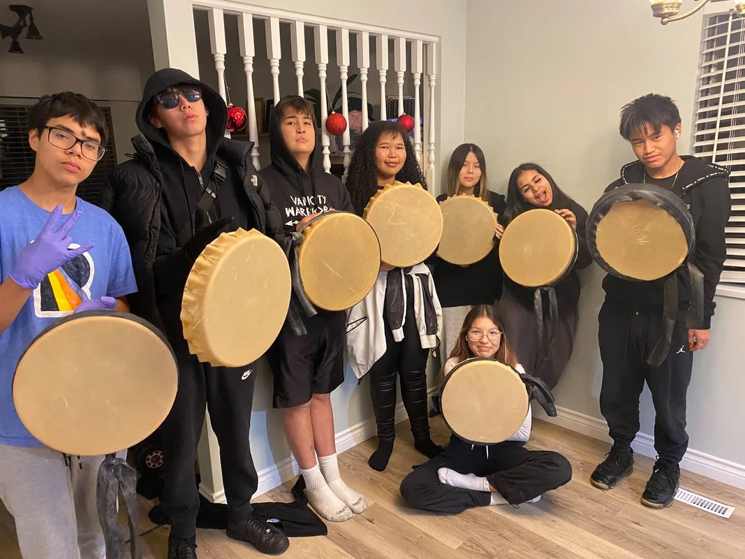 Group of nine young people indoors, holding round, flat, beige objects with black edges. Some are standing, one is sitting on the floor, and one is making a peace sign. They appear excited or playful.
