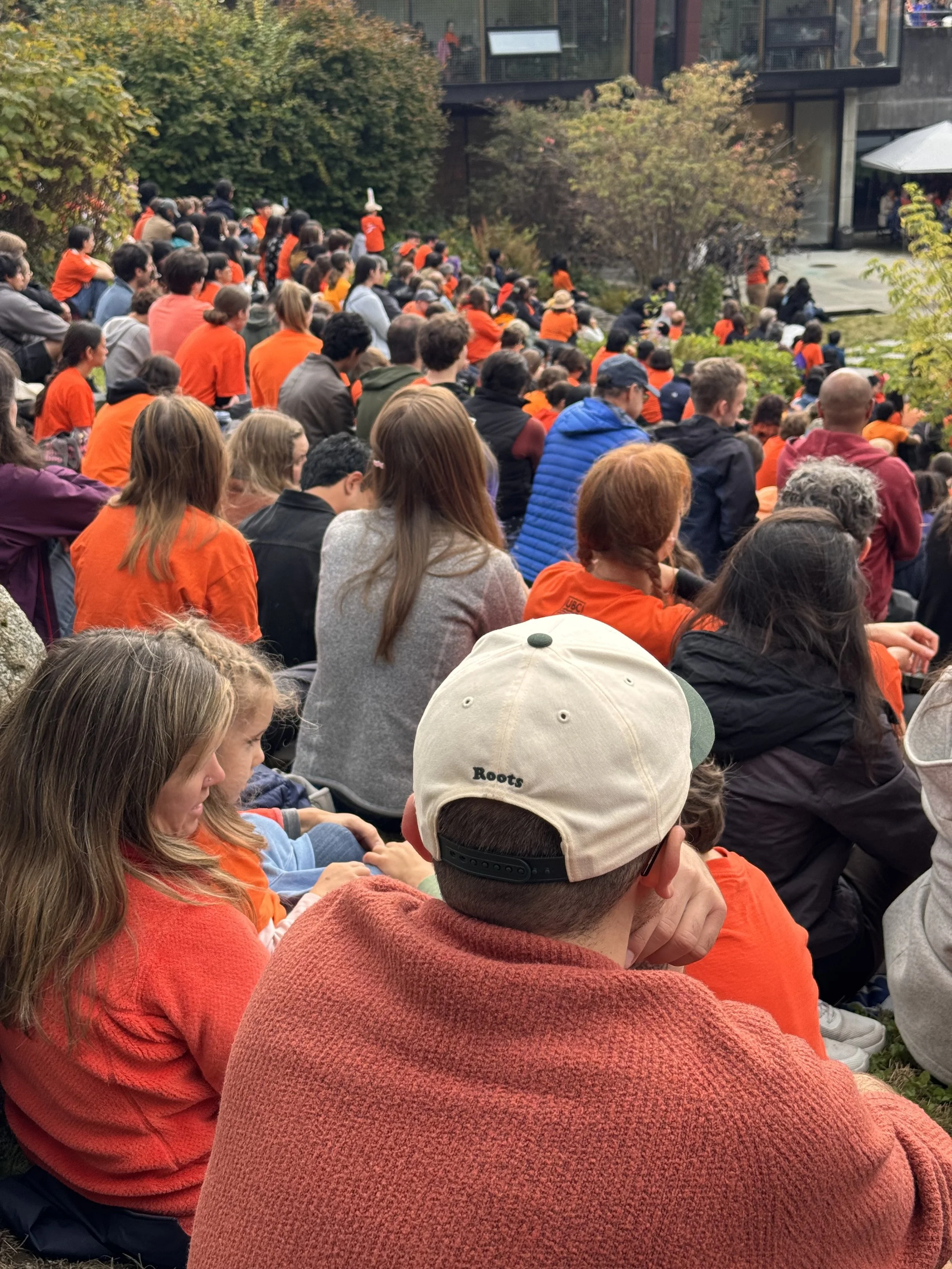 A large crowd of people sitting outdoors, many wearing orange clothing, watching a performance or event in front of a building with glass windows and greenery around.