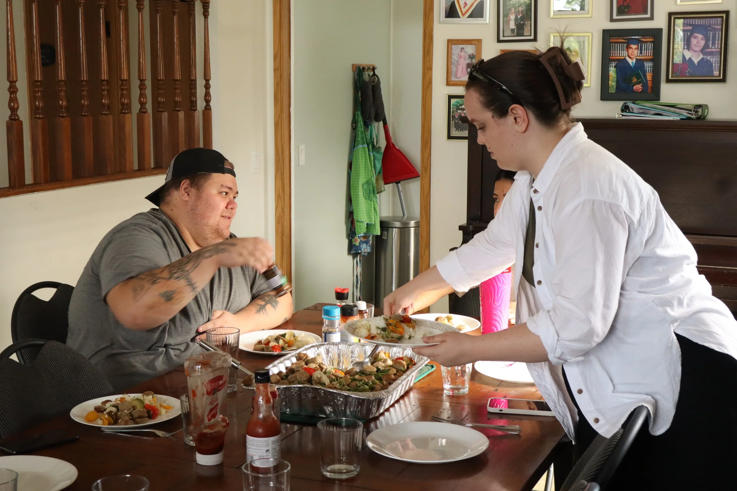 People gathered around a dining table with food, with one person serving a plate to another, in a room decorated with framed photos on the wall.
