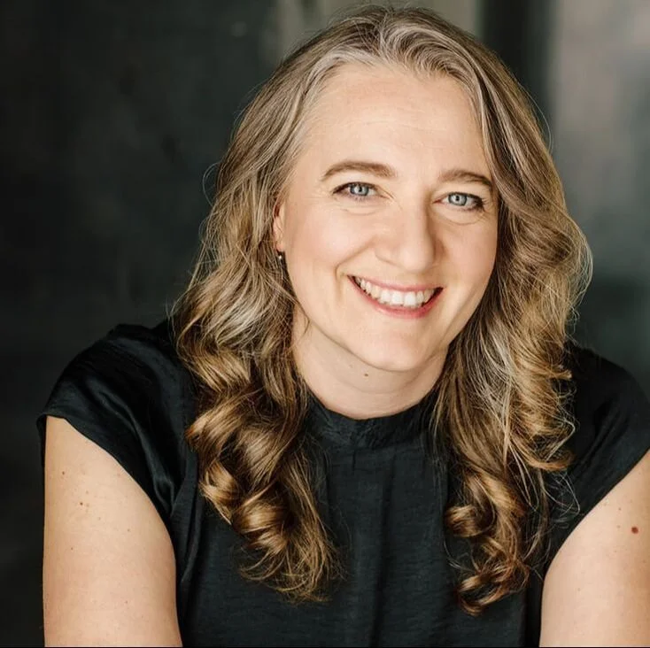 Portrait of a smiling woman with long wavy blonde hair, wearing a black top, against a dark background.