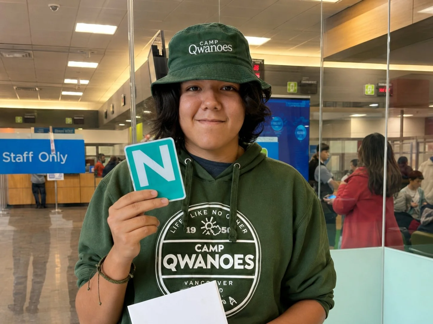 A person holding a green tile with the letter 'N' at an airport terminal, wearing a green hoodie with a "Camp Qwanoes" logo and a matching bucket hat, with a "Staff Only" sign in the background.