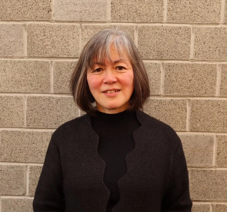 A woman standing in front of a beige brick wall, wearing a black sweater and smiling.