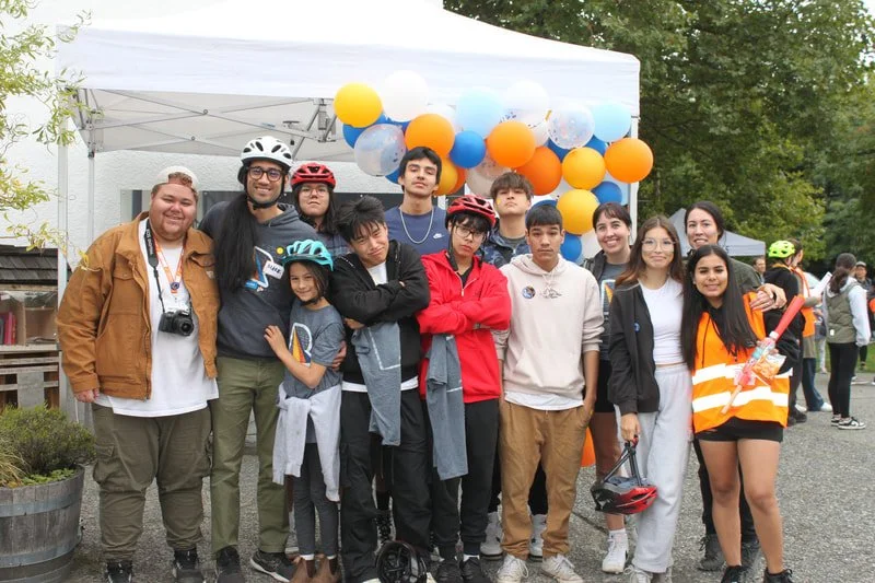 Group of people, mostly teenagers, posing outdoors in front of a white canopy decorated with orange, blue, yellow, and white balloons, some of whom are wearing helmets and casual clothing.