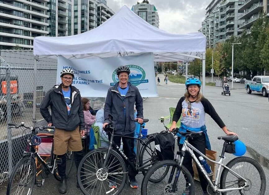Three people standing with bicycles in front of a charity tent on a city street, wearing helmets and casual outdoor clothing.