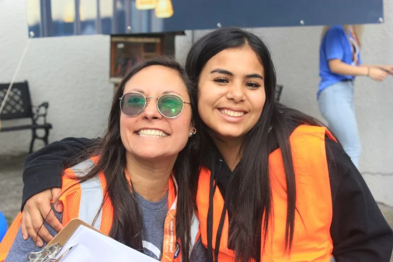 Two smiling women wearing orange safety vests, posing for a photo outdoors.