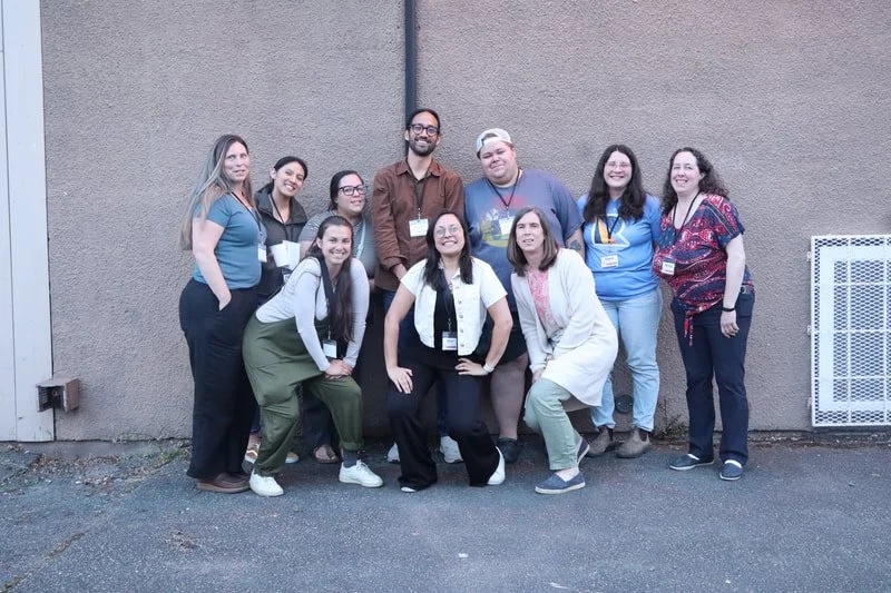 Group of ten diverse people standing and kneeling outside in front of a beige wall, smiling, wearing casual clothing and conference badges.