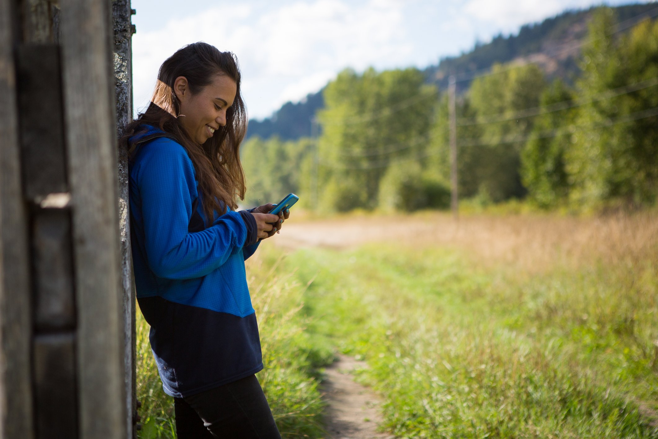 A young woman in a blue jacket smiling and looking at her phone while leaning against a wooden structure outdoors in a green, mountainous area.