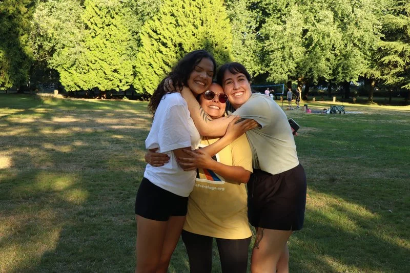 Three young women hugging and smiling in a park with green trees in the background.