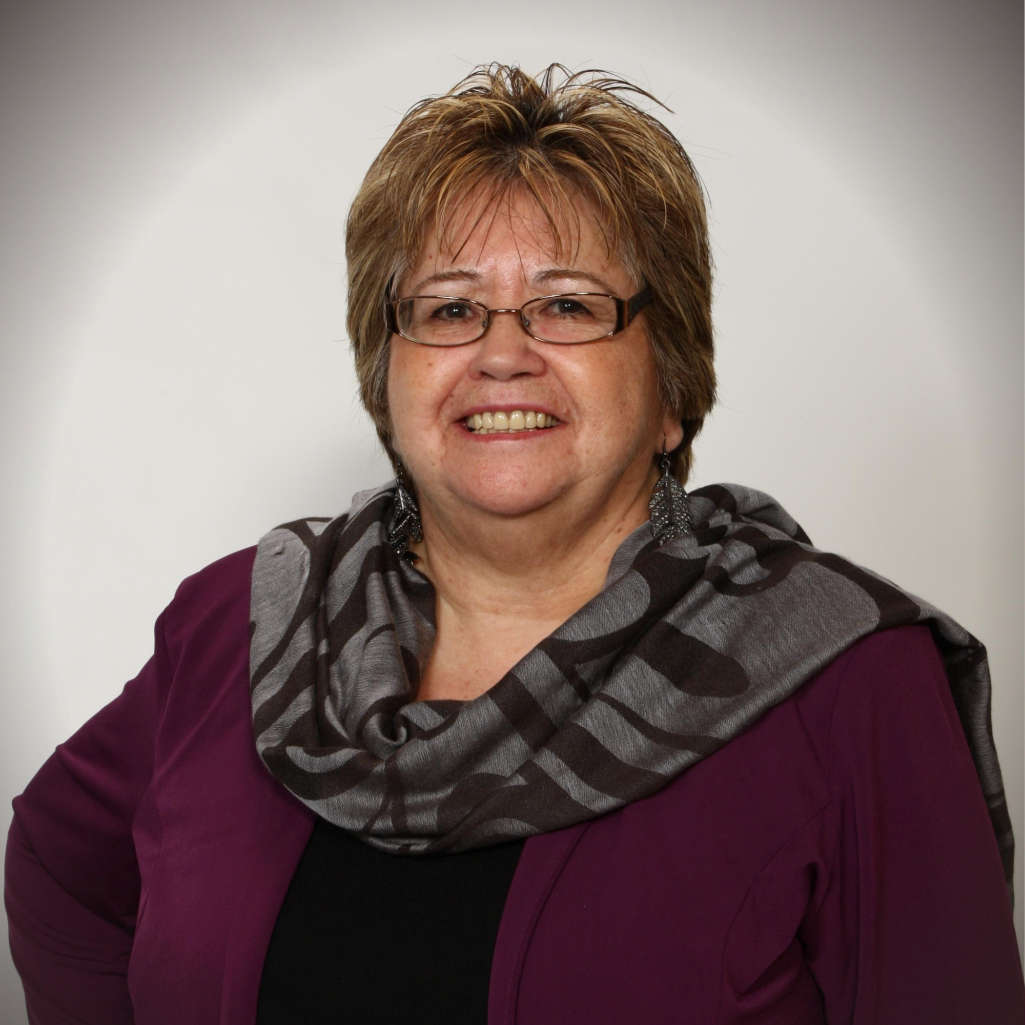 A woman with short brown hair, glasses, and earrings smiling, wearing a striped gray and black scarf, a maroon jacket, and a black top against a plain gray background.