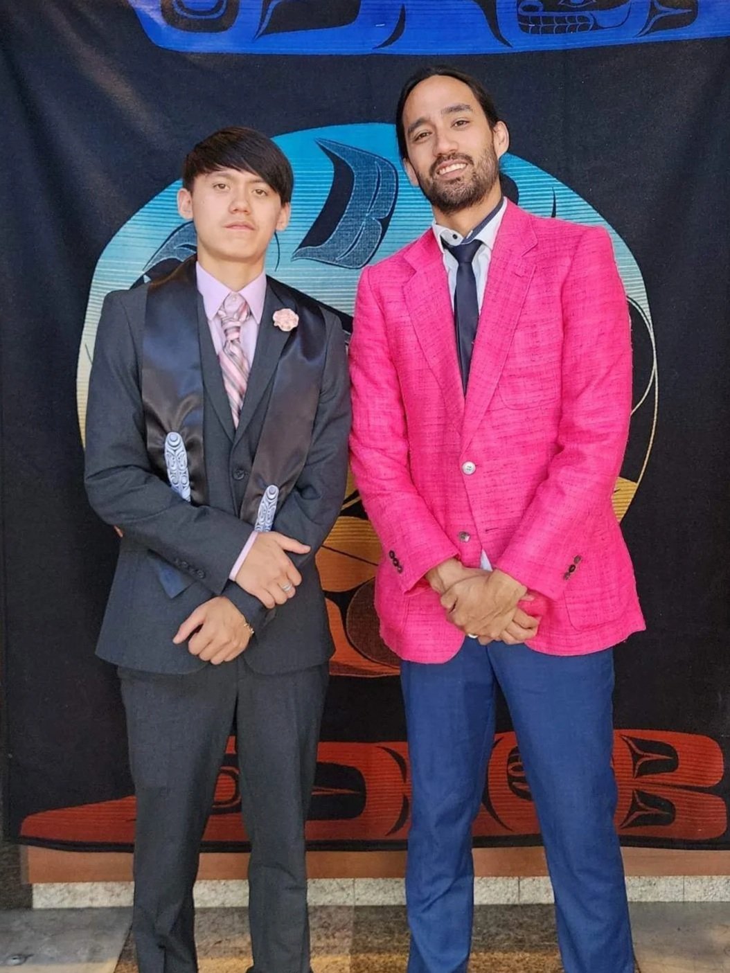 Two men dressed in formal attire standing in front of a decorative banner, one in a dark suit with a floral lapel pin and the other in a bright pink blazer. They are posing for a photo.