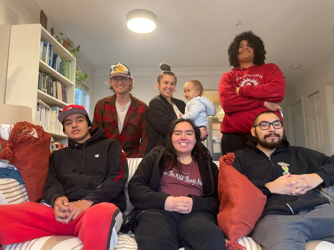 Six people gathered in a living room, including three sitting on the couch and three standing behind them, with a young child in one person's arms. The room has a bookshelf filled with books and a ceiling light.