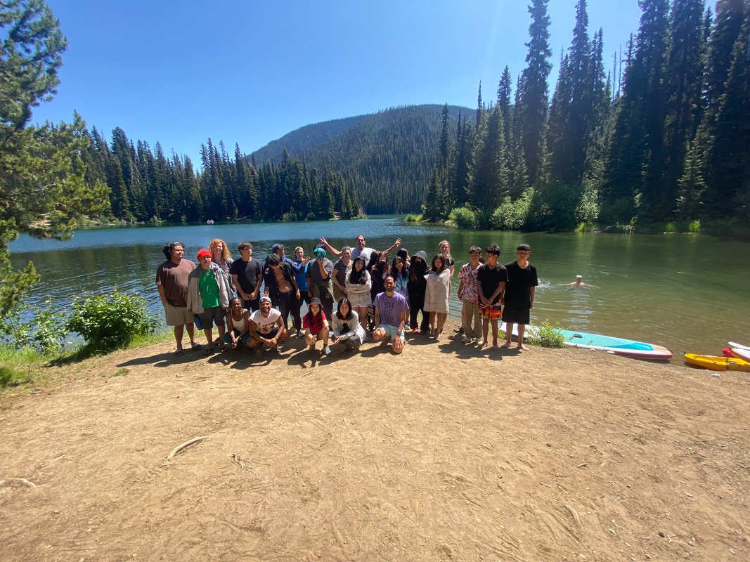 A group of people on a sandy shore near a lake, surrounded by pine trees and mountains, with paddleboards on the water in a forested area on a sunny day.