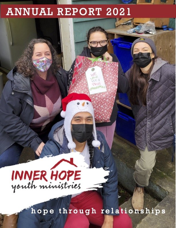 Four women wearing masks are standing together outdoors, smiling for the camera. One woman is holding a large gift bag, and one is wearing a festive snowman hat. There is a sign on the image that reads 'INNER HOPE youth ministries' with a house logo and the tagline 'hope through relationships'. The background shows a blue recycling bin and some wooden shelves.