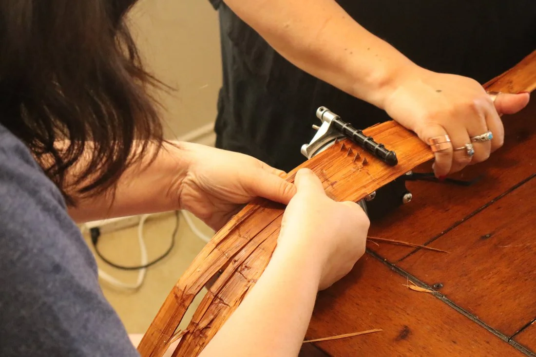 Two women repairing a wooden chair seat, using a staple gun on the wood.