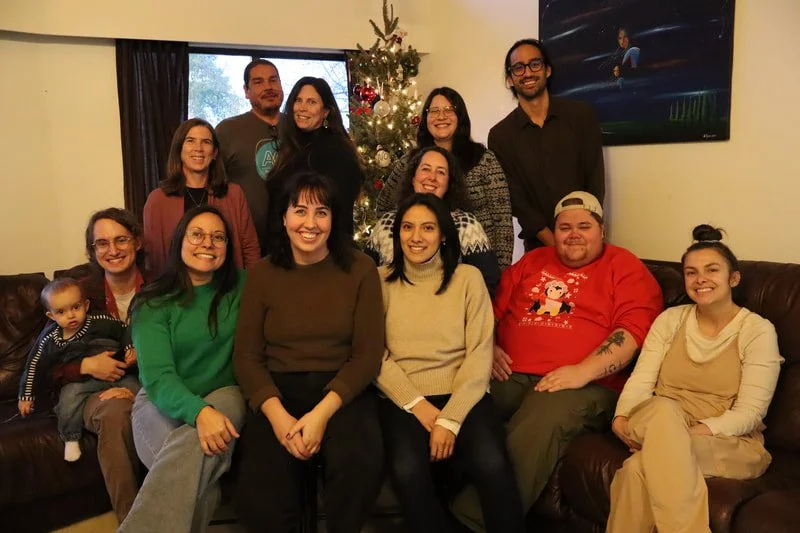 A group of thirteen people, including a child, gathered in a living room during Christmas. They are sitting and standing around a decorated Christmas tree with lights and ornaments. There is a large window behind them and a television on the wall.