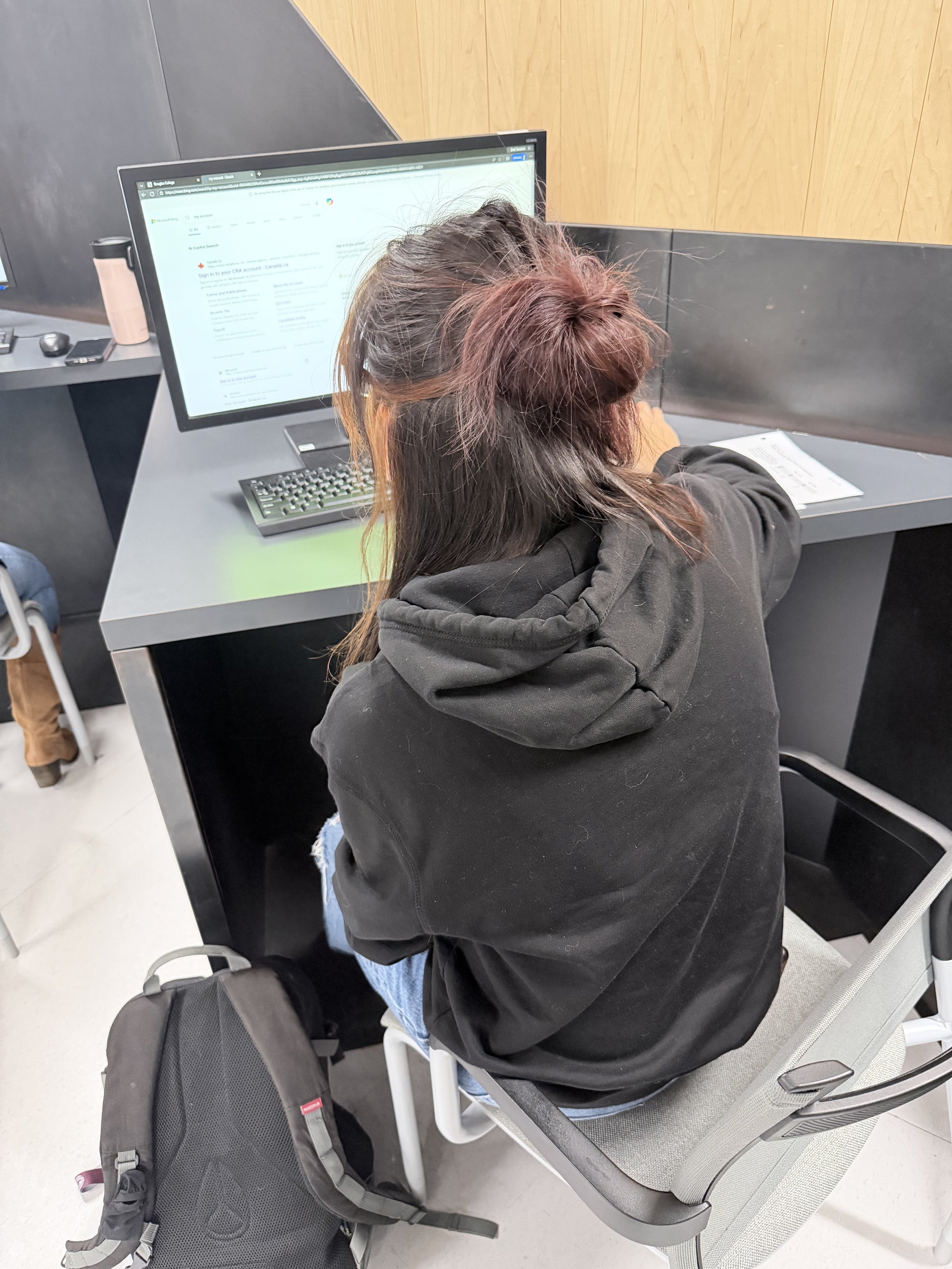 A woman withBraided hair in a bun, wearing a black hoodie, sitting on a gray chair at a desk, working on a computer in a modern office. A black backpack is on the floor next to her.